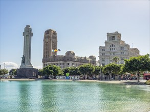 Central square with monument and historic buildings, in front a calm water under a blue sky,