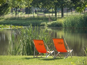 Two orange deckchairs on the shore of a pond in a quiet green park on a sunny day, papenburg,