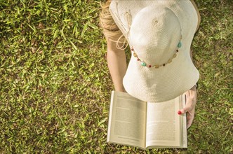 Pretty woman reading book lying on the lawn, seen from above with summer hat