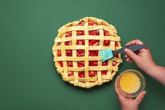 Woman hands washing with egg yolk the pie crust. Top view with strawberries and rhubarb tart.