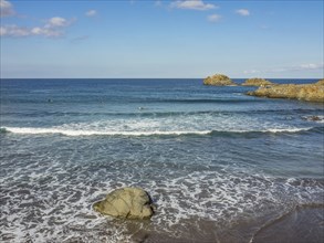 Coastal scene with blue sea and gentle waves, some rocks on and in the water, blue sky with few