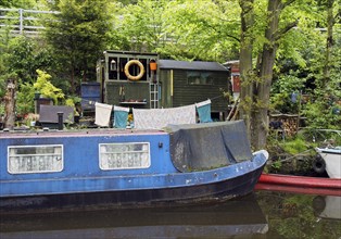 An old scruffy blue narrow boat moored next to wooden sheds with washing on the line on the