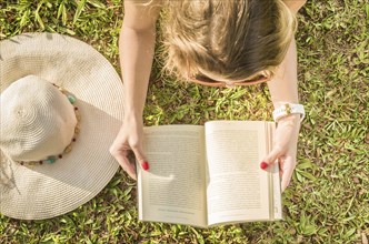 Pretty woman reading book lying on the lawn, seen from above with summer hat