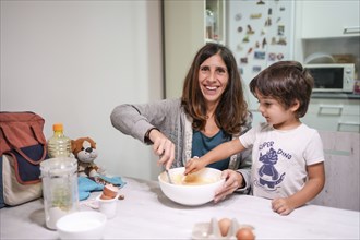 A woman and a child are making a cake together. The woman is smiling and the child is wearing a