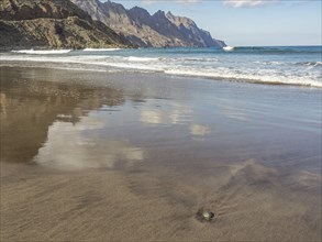 Tidal beach with surrounding mountains and calm sea, waves, clouds reflected in the wet sand,