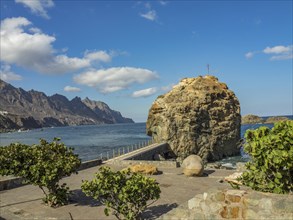 Large rock in the sea, connected by a promenade, surrounded by coastal landscape and blue sky,