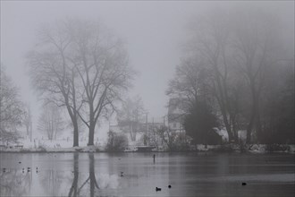 In foggy weather a lake with a house and trees in the background