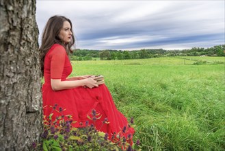 Beautiful woman, in an elegant red dress, sitting on a bench under a tree, with a book and pocket