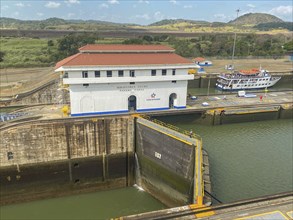 A picture of a lock with a large white building and a small boat. The surroundings are green and