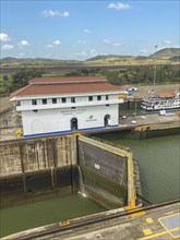 A canal lock next to a large white building. The surroundings consist of green hills and trees