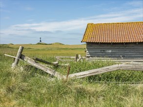 An old wooden barn with a dilapidated wooden fence in front of a blue sky and grassy fields,