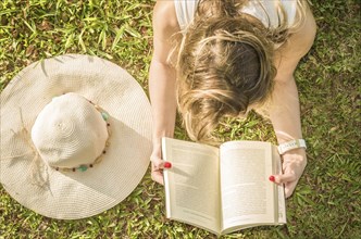 Pretty woman reading book lying on the lawn, seen from above with summer hat