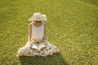 Pretty woman reading book sitting on the lawn, seen from above with summer hat