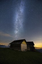 A mountain hut under a clear starry sky with the Milky Way visible and mountains in the background,