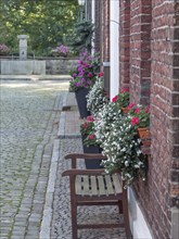 Wooden bench and potted plants with flowers in front of brick houses, along a cobbled alley,