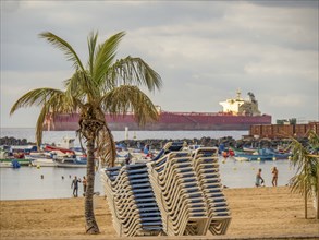 Sun loungers piled up on the beach, in the background a container ship and boats in the water,