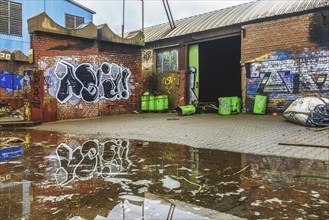 An abandoned courtyard with graffiti, puddles and green rubbish containers next to a brick wall,