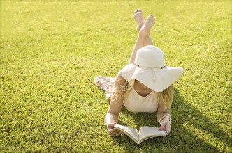 Pretty woman reading book lying on the lawn, seen from above with summer hat