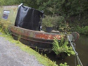 An old rusting houseboat overgrown with weeds moored on the rochdale canal listing to one side and