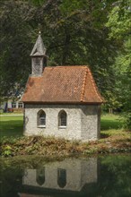 A small stone house with a red tiled roof stands on the edge of a pond, surrounded by dense forest,