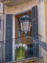 Window with black blinds and flowers on a balcony, above an ornate lantern, palma de Majorca,