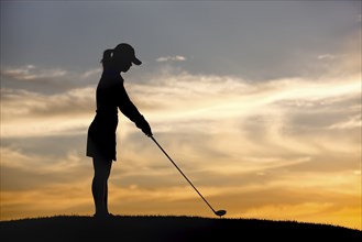 A silhouette of a woman golfer at sunset teeing up on the ball