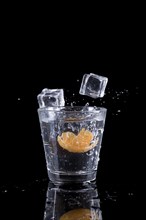 A studio photo of ice cubes being dropped into a glass of water with an orange slice