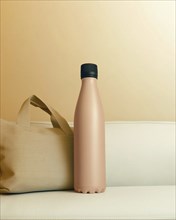 A pink water bottle with a black cap stands next to a beige bag on a beige couch with soft lighting