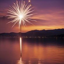 Sparkler glowing at sunset over a lake, reflecting on the water with mountains in the background,