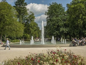 Central fountain in the park with walkers and lush greenery under a blue sky, Bad Lippspringe,