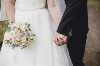 Bride in white dress is holding wedding bouquet. Groom hugs bride. Blurred background.