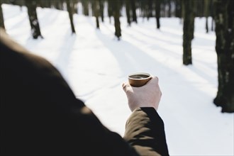 The guy holds a paper cup of coffee in his hand in the snowy forest.