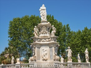 Historic fountain with several statues, surrounded by vegetation and palm trees under a bright blue