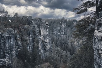 Rugged rocks at Basteibridge during snowfall. Wide view over trees and mountains. National park in