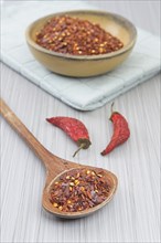 Studio photo of red pepper flakes on a wooden spoon with a yellow wooden bowl of pepper powder and