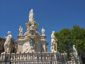 Historic fountain with several statues, surrounded by a railing and vegetation under a blue sky,