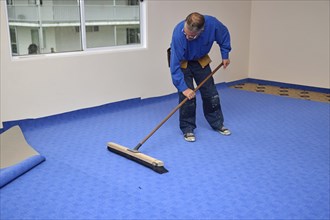 A floorlayer carefully uses a wide broom to smooth out the wrinkles in his newly laid carpet