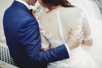 Groom hugs bride back in white dress. Blurred background.