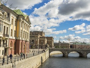 Waterfront promenade of a city with majestic buildings and a bridge over a river, crowded with