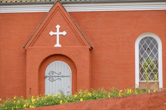 Orange-coloured church facade with white cross and flowers in the foreground, peaceful atmosphere,