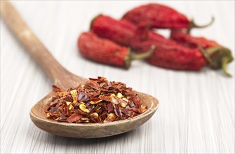 A close up photo of red pepper flakes on a wooden spoon and whole red peppers in the background