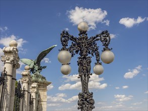 Wrought iron street lamp with ball lamps in front of a blue sky and a winged statue, budapest,