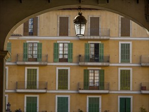 Yellow residential building with green shutters and balconies, framed by an arch with a hanging