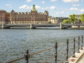 Riverside with a wrought iron railing, a bridge and historic buildings under a sunny sky,