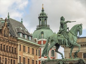 Bronze statue of a rider on a horse in front of a building with a central tower and clear sky,