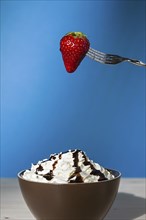 Strawberry suspended from a fork over a cup of whipped cream and chocolate with blue background