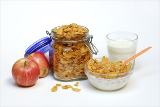 Glass jar and glass bowl with cornflakes, apples, glass with milk, spoon, preserving jar, bowl