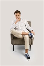 Pensive young handsome man in beige trousers and white shirt sits in armchair in studio