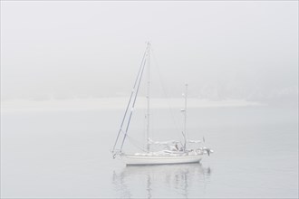 Sailboat, sailing boat, yacht with lowered sails anchored near beach during bad visibility due to