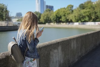 Side view of young girl with backpack reading message from phone while walking along the river